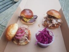 Image of three sandwiches in a box. A sliced brisket slider topped with Lynton gold mustard BBQ sauce and cucumber pickle, a pulled chicken sandwich topped with crispy chicken skin and Lynmouth white sauce, and our pulled pork chicken sandwich with Barbrook BBQ rum sauce and topped with pickled red onion. All sandwiches served on a homemade brioche bun. There is a pot of pickled cabbage in the box as well.